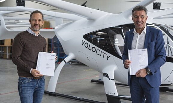 Zwei Männer stehen vor einem helikopterähnlichen Flugobjekt und halten jeweils ein Dokument in der Hand. Der Mann auf der linken Seite hat kurze blonde Haare und trägt einen braunen Pulli, der Mann auf der rechten Seite hat kurze graue Haare und trägt einen blauen Anzug.