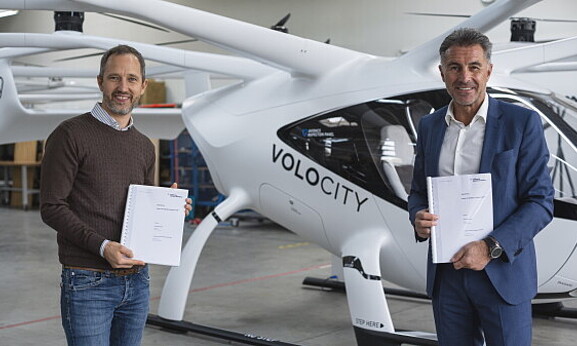 Two men are standing in front of a helicopter-like flying object, each holding a document in their hands. The man on the left has short blond hair and is wearing a brown sweater, while the man on the right has short gray hair and is wearing a blue suit.