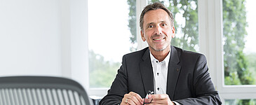 Lächelner Mann mit kurzen grauen Haaren, dunkelgrauem Sackko und weißem Hemd sitzt an einem Tisch, hält eine dunkel umrandete Brille in der Hand und schaut in die Kamera