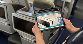 Ein Mann steht am Sitzplatz in der Flugzeugkabine und hält ein Tablet in den Händen. Auf dem Tablet sieht man den Sitzplatz wie durch eine Kamera.