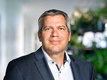 Mann mit kurzen grauen Haaren, braunen Augen, dunkelgrauem Jackett und weißem Hemd